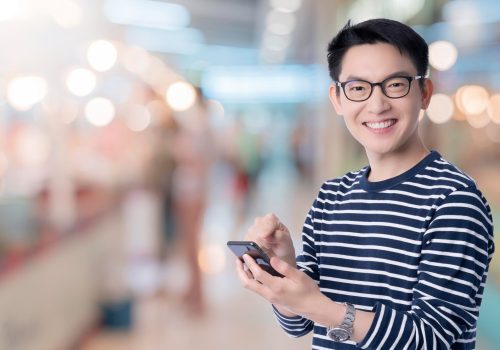 smart-casual-asian-happiness-male-wear-glasses-smile-cheerful-hand-use-smartphone-ready-press-buy-button-with-shopping-mall-abstract-blur-background-technology-communication-ideas-concept